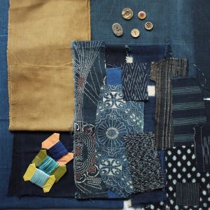 Op de afbeelding: Een verzameling stofstalen met diverse indigoblauwe patronen, waaronder geometrische, florale en gestreepte ontwerpen, naast een effen beige stofstuk, vier kleine ronde knopen en drie klossen met blauw en groen borduurgaren.