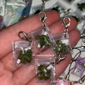 Puede incluir: Una mano sosteniendo cuatro peque&ntilde;as bolsas de pl&aacute;stico transparentes con cierres plateados. Cada bolsa contiene una peque&ntilde;a cantidad de material verde frondoso. Las bolsas est&aacute;n unidas a cadenas plateadas. El texto "@creationsbylace" es visible en la imagen.