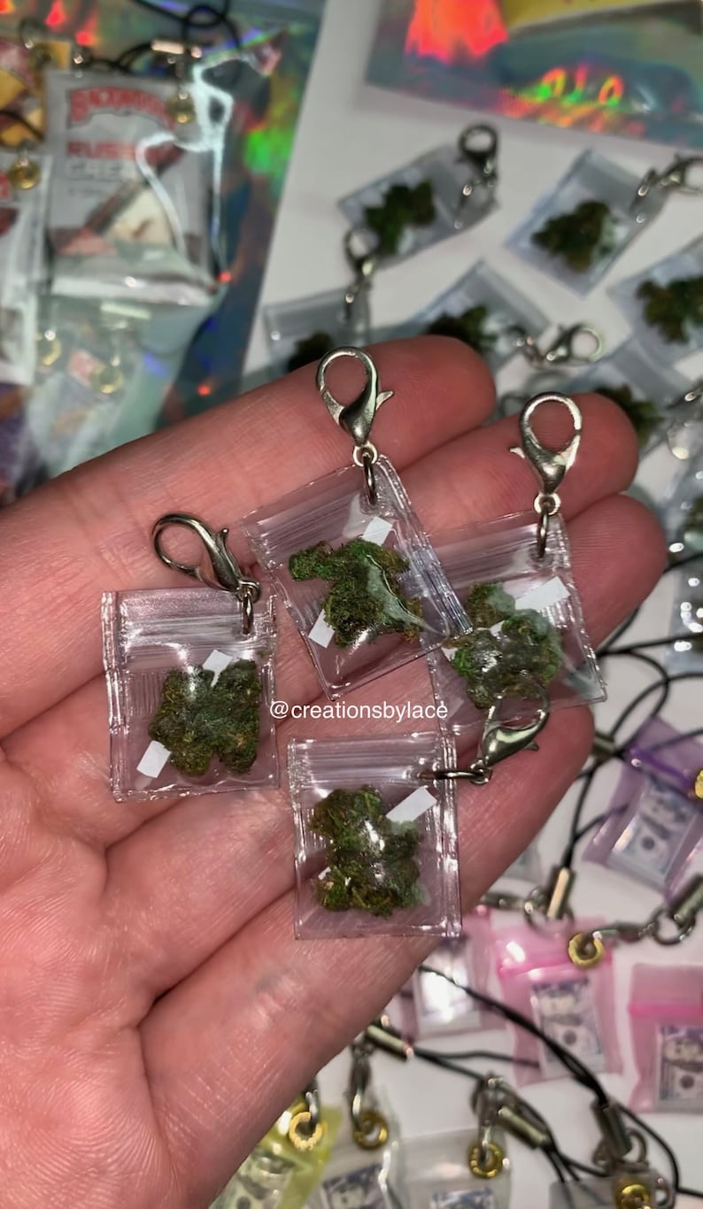 Puede incluir: Una mano sosteniendo cuatro peque&ntilde;as bolsas de pl&aacute;stico transparentes con cierres plateados. Cada bolsa contiene una peque&ntilde;a cantidad de material verde frondoso. Las bolsas est&aacute;n unidas a cadenas plateadas. El texto "@creationsbylace" es visible en la imagen.