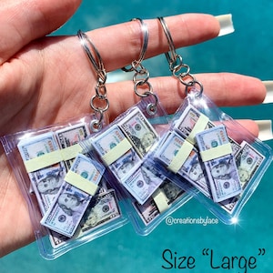 May include: Three clear plastic keychain charms with stacks of fake money inside. The charms are attached to silver key rings. The text "Size "Large"" is at the bottom of the image.