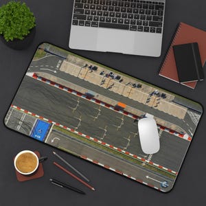May include: A black mousepad featuring a detailed aerial view of a race track with red and white barriers. The mousepad has a black border and a white computer mouse is on the track. A laptop, coffee cup, and pens are also visible.