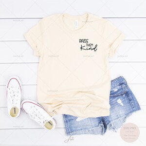 Peut inclure: Un t-shirt beige avec le texte "Raise them kind" en lettres noires. Le t-shirt est posé sur une surface en bois blanc avec une paire de baskets blanches et un short en jean bleu.