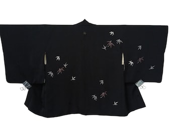May include: A black haori jacket featuring wide sleeves and an open front. The garment is decorated with scattered, embroidered motifs of bamboo leaves in shades of white and muted reddish-brown. Two silver metal binder clips are attached to the sides.