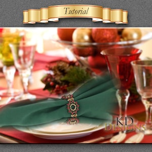May include: A green napkin ring with red and gold beads. The napkin ring is on a green napkin. The napkin is on a white plate. The plate is on a red placemat. There are glasses and a bowl of red and gold ornaments in the background. The text "KD DESIGNS" is visible in the bottom right corner of the image.
