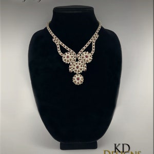 Peut inclure: Un collier en argent avec un motif en rubis et en cristal. Le collier est sur un buste de mannequin noir. Le texte "Ruby Elegance" est au-dessus du collier. Le texte "KD DESIGNS" est en dessous du collier.