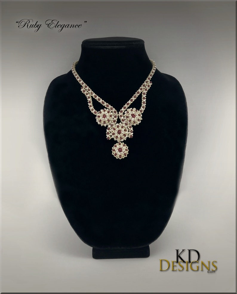 Peut inclure: Un collier en argent avec un motif en rubis et en cristal. Le collier est sur un buste de mannequin noir. Le texte "Ruby Elegance" est au-dessus du collier. Le texte "KD DESIGNS" est en dessous du collier.