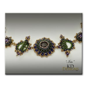 Puede incluir: Primer plano de un collar de cuentas con una piedra preciosa de color p&uacute;rpura oscuro en el centro. El collar est&aacute; hecho con cuentas verdes, doradas y moradas. El texto "Iris" y "KD Designs" es visible en la esquina inferior derecha de la imagen.