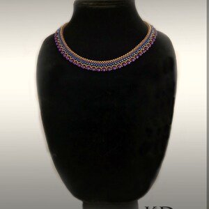 May include: A gold and purple beaded necklace on a black display stand. The necklace has a simple design and is labeled "Simplicity".