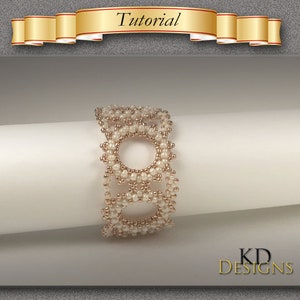 Può includere: Un braccialetto di perline bianche e dorate con un design circolare. Il braccialetto è su una superficie bianca. KD Designs.