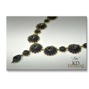 Puede incluir: Un collar de cuentas con un esquema de color verde, azul y dorado. El collar presenta una serie de medallones redondos de cuentas con una piedra preciosa p&uacute;rpura central. El collar est&aacute; etiquetado como "Iris" KD Designs.