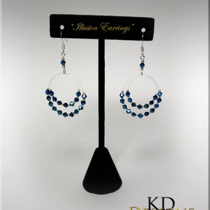 May include: A pair of silver hoop earrings with a double row of dark blue crystal beads. The earrings are displayed on a black stand with the text "Illusion Earrings" above them.