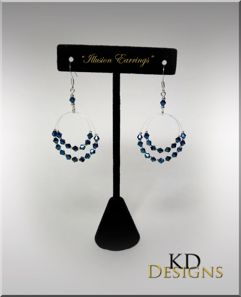 May include: A pair of silver hoop earrings with a double row of dark blue crystal beads. The earrings are displayed on a black stand with the text "Illusion Earrings" above them.