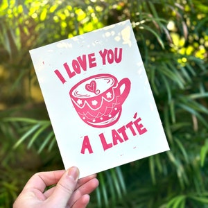 May include: A white card with pink lettering that reads "I love you a latte" and a pink illustration of a coffee cup with a heart in the coffee.