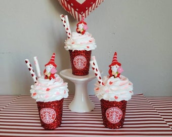 May include: Three red glitter Starbucks coffee cups with white whipped cream, pink hearts, and a small gnome figurine on top. The cups are on a white pedestal and a red and white striped surface.