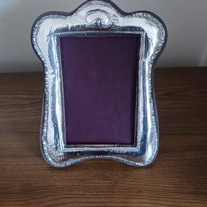 Puede incluir: Un marco de fotos plateado con un borde festoneado decorativo. El marco tiene un interior morado oscuro y se apoya sobre una superficie de madera. El diseño del marco incluye un pequeño detalle ornamentado en la parte superior.