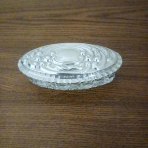 Könnte beinhalten: Eine ovale, silberfarbene Schmuckschatulle mit einem geprägten Deckel. Der Deckel hat ein dekoratives Design mit Wirbelmustern und einem zentralen Oval. Der Boden besteht aus klarem, facettiertem Glas. Die Schatulle steht auf einer Holzoberfläche.