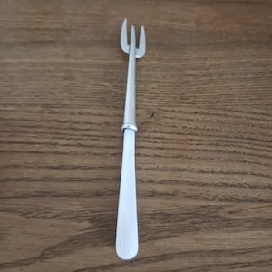 May include: A three-pronged silver-toned metal cocktail fork featuring a smooth, solid white handle. The utensil has a slender neck that connects the metallic tines to the opaque white grip, creating a clean and functional design for serving small bites.