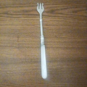 Pode incluir: Um garfo de coquetel de metal prateado com uma cabeça de três pontas e uma seção de pescoço de metal trançado. O cabo é composto por um material branco opaco e liso, proporcionando um contraste nítido com a parte superior metálica do utensílio.