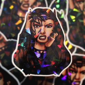 Puede incluir: Una pegatina holográfica con una mujer de cabello oscuro y cuernos, su rostro parcialmente oscurecido por un diseño abstracto brillante. Tiene una expresión de angustia y se está sujetando la cabeza con una mano.