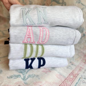 May include: A stack of four gray sweatshirts with embroidered initials in various colors: light blue, pink, green, and navy. The initials are large and centered on the front of each sweatshirt, creating a personalized look.