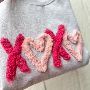 May include: A gray sweatshirt with the letters "XOX" in a textured, fluffy, pink and red design. The letters are in a heart shape. The sweatshirt is a casual, comfortable style.
