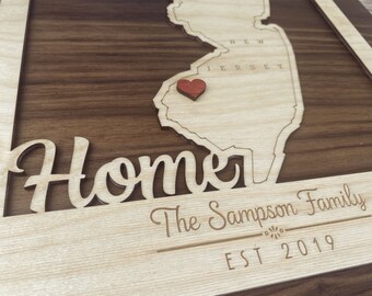 May include: A wooden sign featuring a cutout of the state of New Jersey, a red heart in the centre, and the text "Home" in a decorative font. Below the state is the text "The Sampson Family EST 2019".