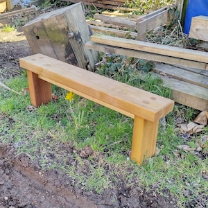 May include: A simple wooden bench with a natural finish. The bench is made of solid wood and has a rectangular shape. It is sitting on a grassy area with dirt.