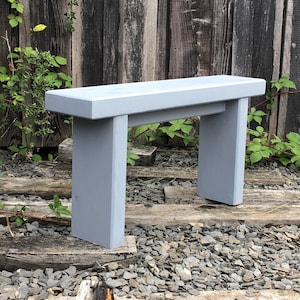 May include: A light grey wooden bench with a simple design. The bench is made of solid wood and has a smooth finish. It is sitting on a bed of grey gravel.