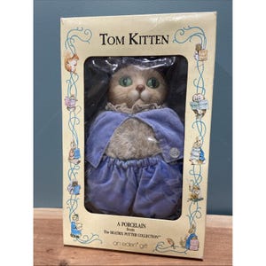 May include: A porcelain Tom Kitten doll from The Beatrix Potter Collection, presented in a decorative box. The doll is dressed in a blue outfit with a ruffled collar and has green eyes. The box features illustrations of other Beatrix Potter characters.