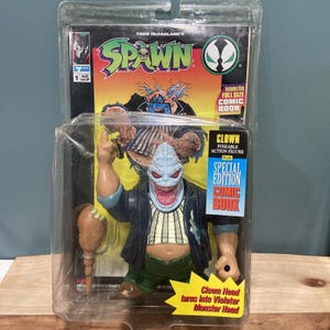 May include: A Todd McFarlane Spawn Clown action figure in original blister packaging. The figure has a pale, textured head, a dark jacket, and green pants, holding a brown club. The packaging includes a full-size comic book and text stating the clown head turns into a Violator monster head.