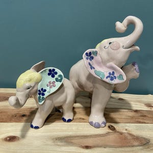 May include: Two ceramic elephant figurines. The elephants are light pink with blue and pink floral designs on their ears. One elephant has its trunk raised. The figurines are on a wooden surface.