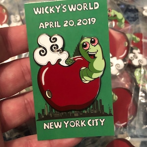 May include: A green enamel pin with a red apple, a green worm with red eyes, and white smoke. The pin says "Wicky's World", "April 20, 2019", and "New York City".