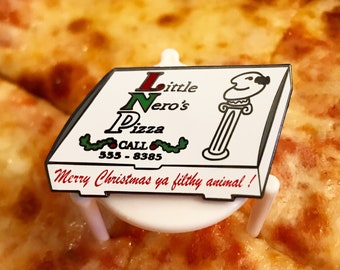 May include: A white and red pizza box pin with the text "Little Nero's Pizza" and "Call 555-8385". The pin has a green holly design and the text "Merry Christmas ya filthy animal!" on the bottom.