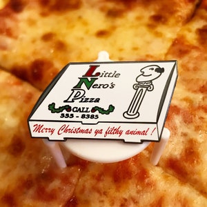 Könnte beinhalten: Eine weiße und rote Pizzakartonstecknadel mit dem Text "Little Nero's Pizza" und "Call 555-8385". Die Stecknadel hat ein grünes Stechpalmen-Design und den Text "Merry Christmas ya filthy animal!" unten.