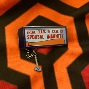 Puede incluir: Un pin de esmalte azul y dorado con fondo blanco. El pin dice "Break Glass in Case of Spousal Insanity" y tiene un pequeño martillo unido a una cadena.