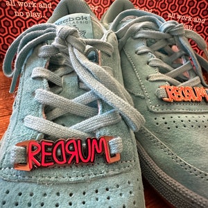 May include: A pair of light blue suede sneakers with a white Reebok logo on the side. The laces are tied and the shoes have a metal charm with the word "REDRUM" written in red letters.