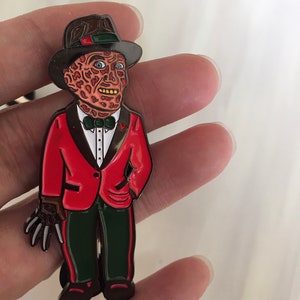 May include: A metal pin featuring a stylized character in a red jacket, green pants, and a brown hat. The character has a scarred face and a clawed glove. The pin is a collectible item, likely inspired by a horror movie.