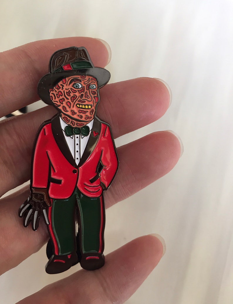 May include: A metal pin featuring a stylized character in a red jacket, green pants, and a brown hat. The character has a scarred face and a clawed glove. The pin is a collectible item, likely inspired by a horror movie.