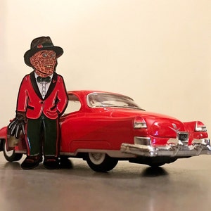 May include: A red enamel pin of a character in a red suit and black hat, leaning against a red 1953 Cadillac toy car. The pin features a character with a burned face, black gloves, and a black bow tie. The car has chrome accents and white tires.