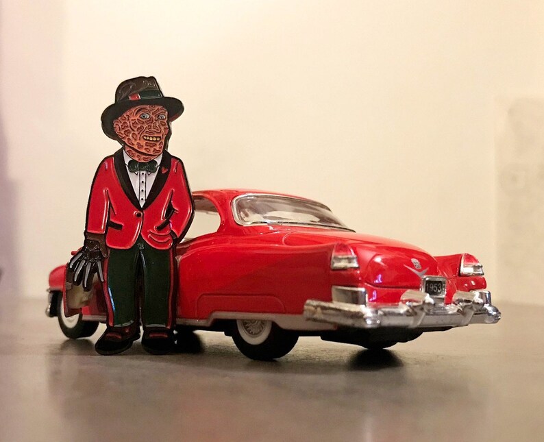 May include: A red enamel pin of a character in a red suit and black hat, leaning against a red 1953 Cadillac toy car. The pin features a character with a burned face, black gloves, and a black bow tie. The car has chrome accents and white tires.