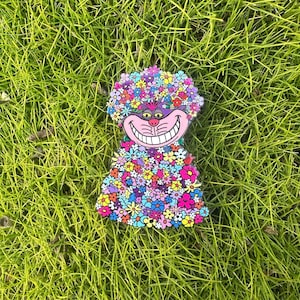 May include: A purple and pink enamel pin of the Cheshire Cat from Alice in Wonderland. The cat is wearing a crown of flowers and has a wide grin.