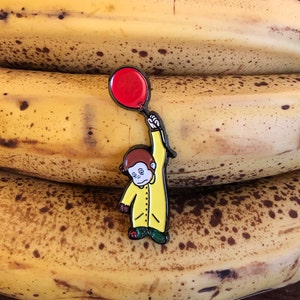 May include: A yellow enamel pin of a monkey wearing a yellow raincoat and holding a red balloon. The pin is on top of a stack of three bananas.