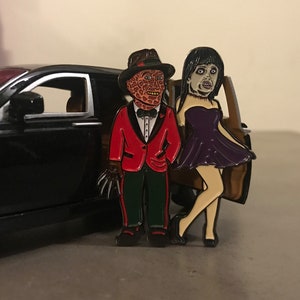May include: Enamel pin set featuring Freddy Krueger in a red tuxedo and a woman in a purple dress. The pin set is next to a black car. The Freddy Krueger pin has a brown hat and a glove with sharp claws.