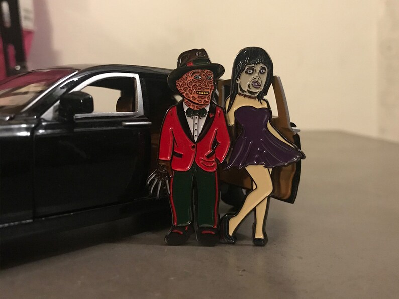 May include: Enamel pin set featuring Freddy Krueger in a red tuxedo and a woman in a purple dress. The pin set is next to a black car. The Freddy Krueger pin has a brown hat and a glove with sharp claws.