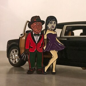 May include: Enamel pin set featuring two horror movie characters. One character is wearing a red blazer, green pants, and a fedora. The other character is wearing a purple dress. The pin set is placed in front of a black toy car.