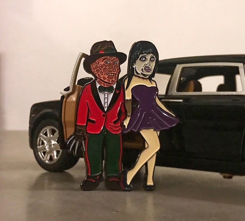 May include: Enamel pin set featuring two horror movie characters. One character is wearing a red blazer, green pants, and a fedora. The other character is wearing a purple dress. The pin set is placed in front of a black toy car.