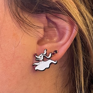 Op de afbeelding: Witte en zwarte Zero-oorstekers. De oorbellen hebben een cartoon spookhond ontwerp met een rode neus en een kleine rode edelsteen. De oorbellen zijn te zien op het oor van een persoon.