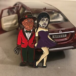 May include: Enamel pin featuring two cartoon characters, one in a red suit and fedora, the other in a purple dress. The pin is set against a backdrop of a red car with an open door.
