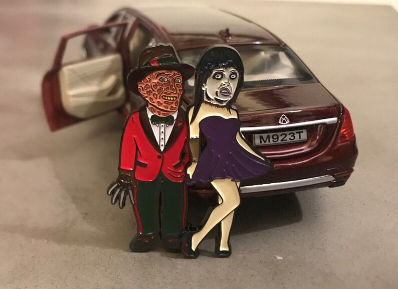May include: Enamel pin featuring two cartoon characters, one in a red suit and fedora, the other in a purple dress. The pin is set against a backdrop of a red car with an open door.