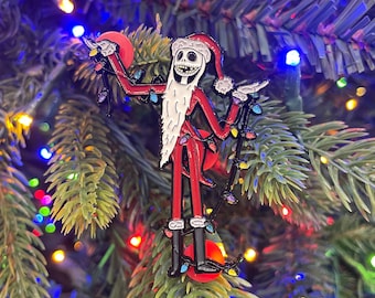 May include: A red and black enamel pin of Jack Skellington dressed as Santa Claus, holding a string of Christmas lights. The pin is attached to a Christmas tree with green needles and colorful lights.